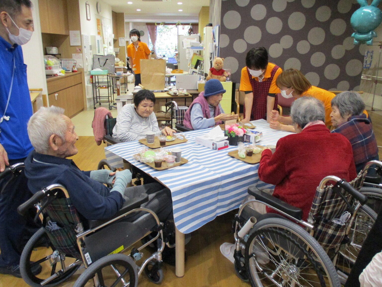 介護施設の入居者がスタッフと共に食事や活動をしている風景。複数の高齢者が車椅子で参加。